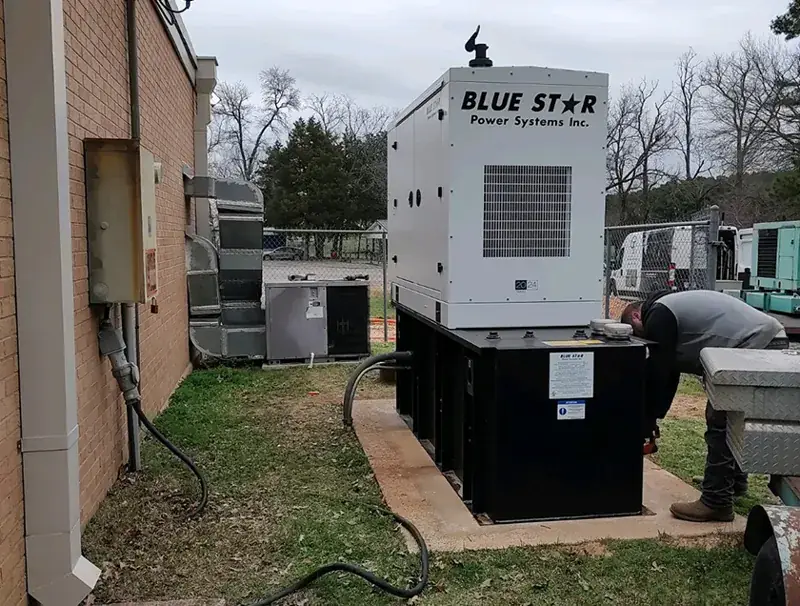 Tall white Blue Star Power Systems generator on a black base on a concrete pad beside a brick building, with a man in a gray vest leaning over the base and black cables on the grass.