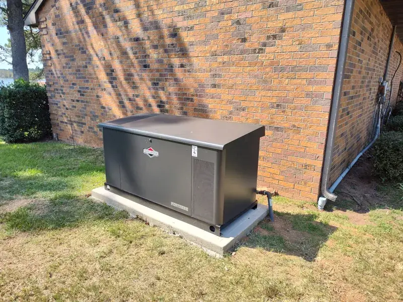 Charcoal-gray Briggs & Stratton Fortress generator on a concrete pad beside a tan-and-brown brick house, with a pipe connection at its right side and water visible through the trees at left.