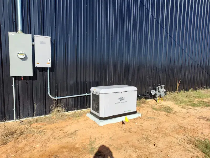 White Briggs & Stratton PowerProtect generator on a pad in red soil beside a dark metal building, with conduit to an electric meter and a gray Briggs & Stratton box, and a gas regulator at right.
