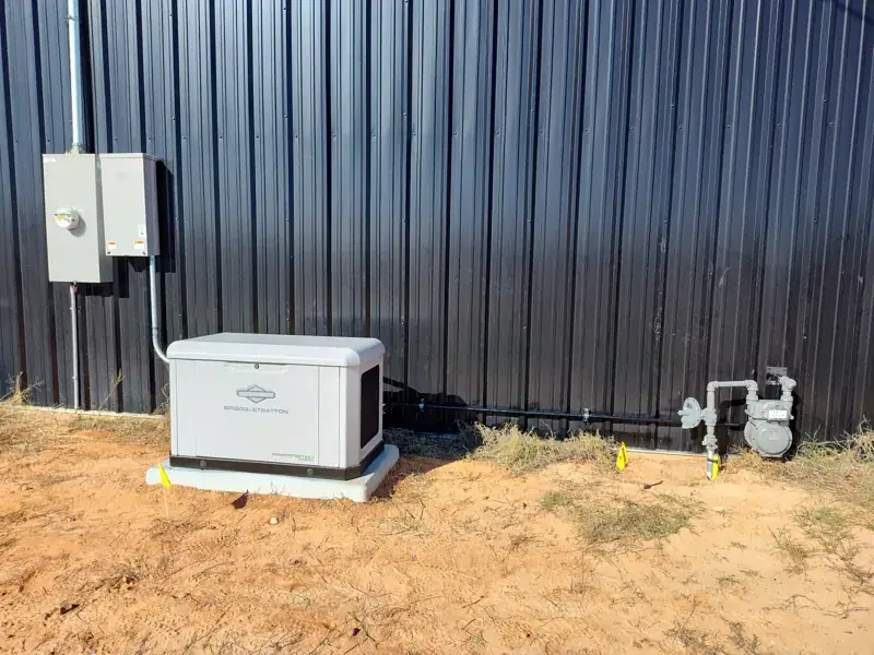 White Briggs & Stratton PowerProtect generator on a pad beside a dark metal building, with a black gas line running along the wall from a gas meter and regulator, and yellow marker flags in red soil.