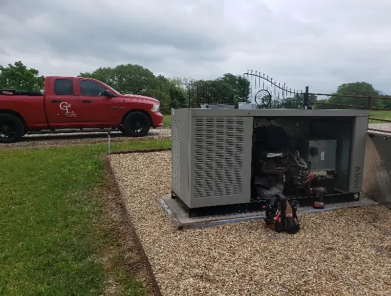 Gray generator in a gravel bed with a side panel removed to show the engine, a drill on its pad and a tool bag in front, a red GenTech pickup on the gravel drive and a metal gate behind.