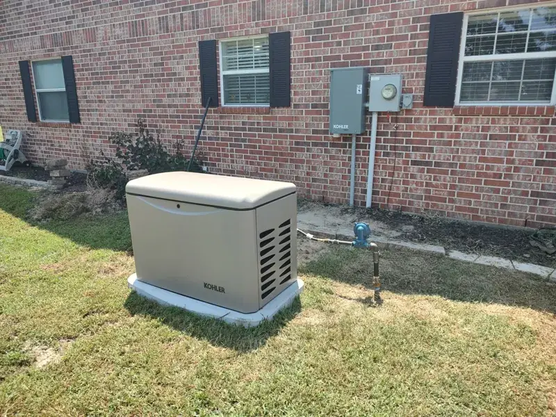 Tan Kohler home standby generator on a white pad beside a red-brick house with black shutters, a gas line connected at its side and a gray Kohler box next to the electric meter.