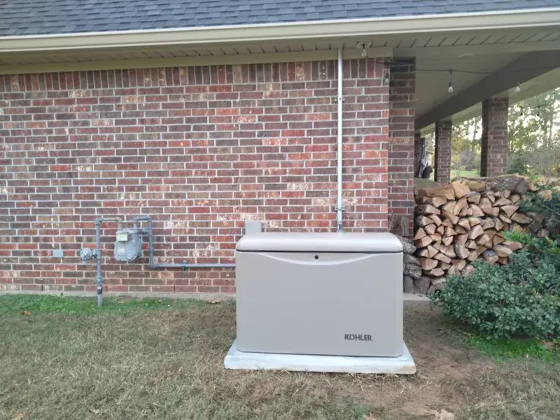 Tan Kohler home standby generator on a pad beside a red-brick house, with a gas meter and gas line on the wall at left and stacked firewood at the edge of a covered patio at right.