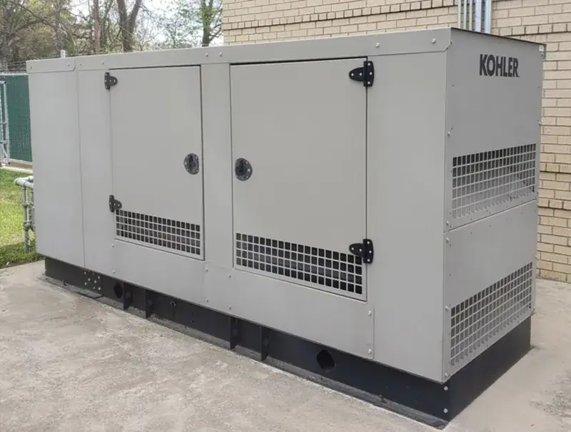 Large gray Kohler generator in a metal enclosure with two hinged access doors and vent grilles, on a concrete pad beside a tan-brick building and a chain-link fence.