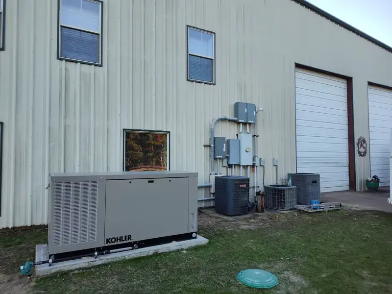 Kohler generator on a concrete pad beside a cream metal building with large garage doors, near gray Kohler electrical boxes, an electric meter and three air-conditioning units.