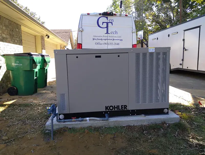 Kohler generator on a concrete pad beside a driveway, with a brick garage at left, a white trailer at right and the GenTech van with its office number behind.