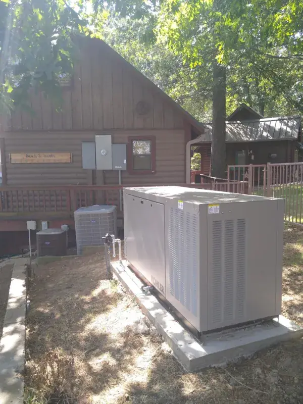 Tan Kohler generator on a long concrete pad under shade trees beside a log-sided home with a wooden deck, two air-conditioning units and gray electrical boxes, one labeled Kohler.