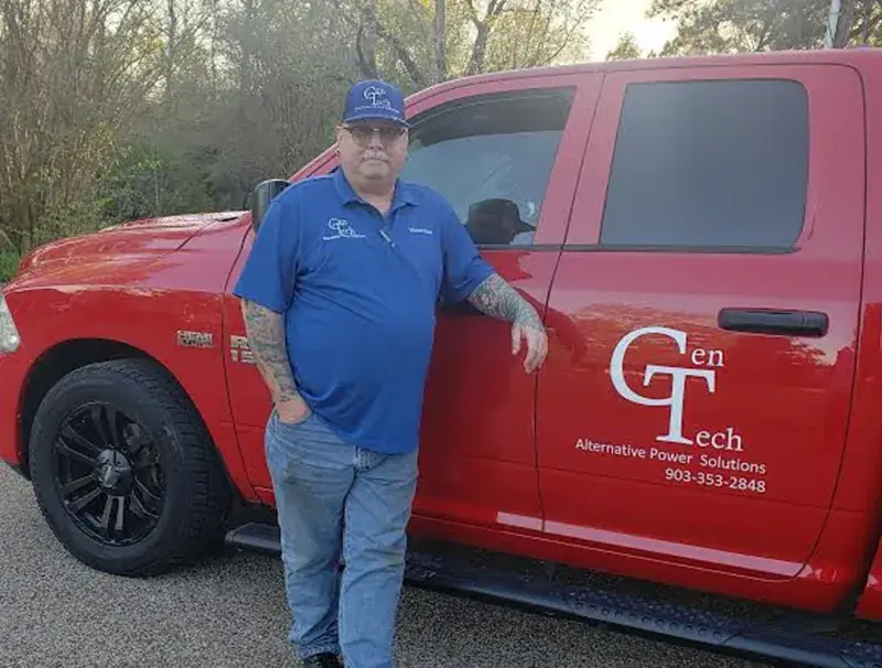 Mike Glisan in a blue GenTech polo and cap leaning on a red pickup lettered GenTech Alternative Power Solutions, 903-353-2848.