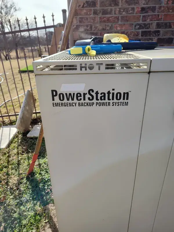 Cream PowerStation Emergency Backup Power System generator beside a brick wall and metal fence.